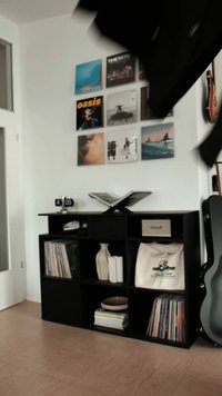 Svart trähylla med öppna fack som innehåller böcker, en vas och vinylskivor. En klocka och en högtalare står på toppen.