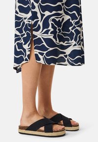 Dunkelblaues Kleid mit weißen Blumenmustern, kombiniert mit schwarzen Sandalen mit Überquerungsgurt und einer geflochtenen Jutearbeit. Seitenschlitz sichtbar.