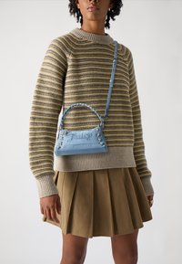 Ανοιχτό μπλε τσάντα χειρός από μαλακό υλικό, με στριμμένο χερούλι, κεντημένο λογότυπο και διακοσμητικά κορδόνια. Συνδυάζεται με μια ριγέ πουλόβερ.