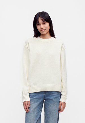 Jeune femme aux cheveux noirs et lisses, portant un pull en maille blanc et un jean bleu, debout devant un fond clair uni.