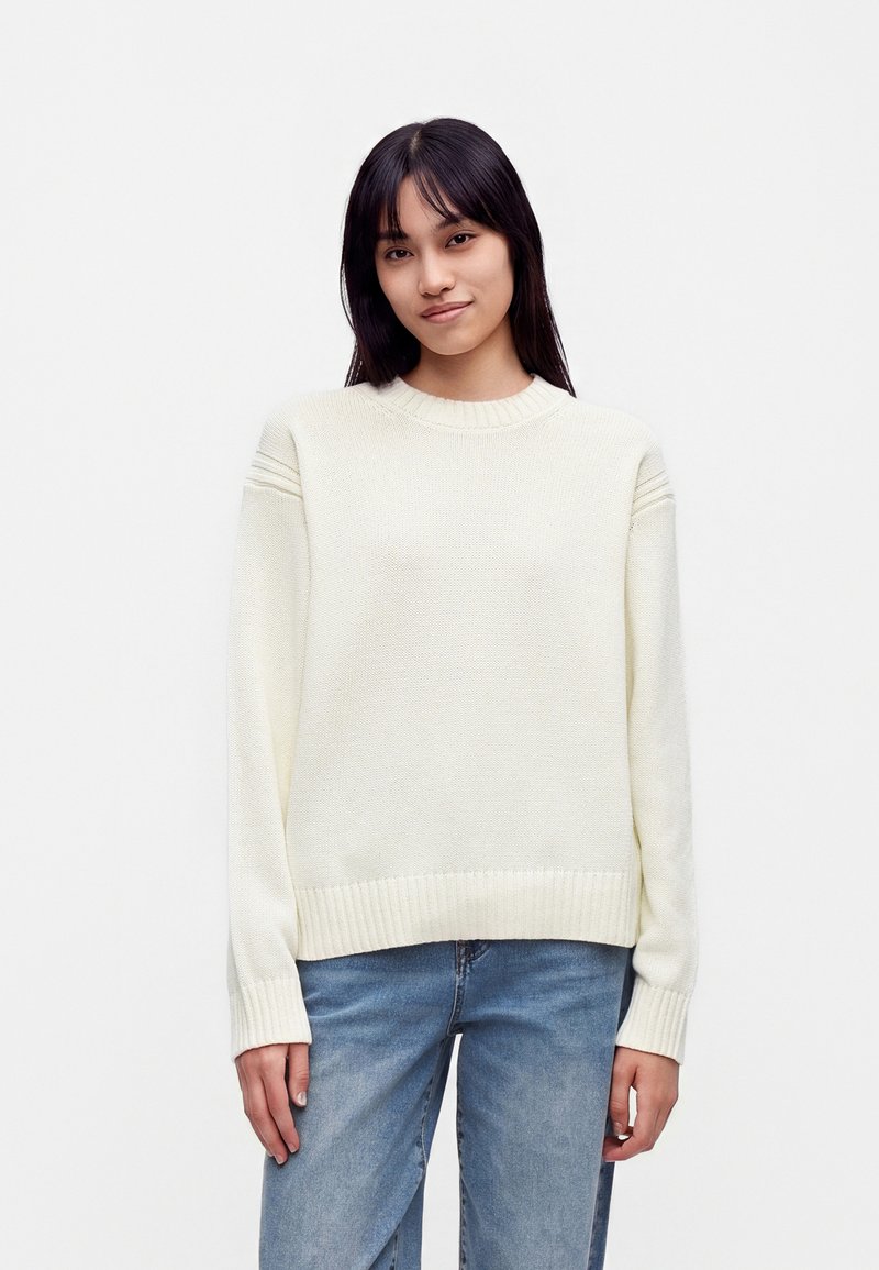 Jeune femme aux cheveux noirs et lisses, portant un pull en maille blanc et un jean bleu, debout devant un fond clair uni.