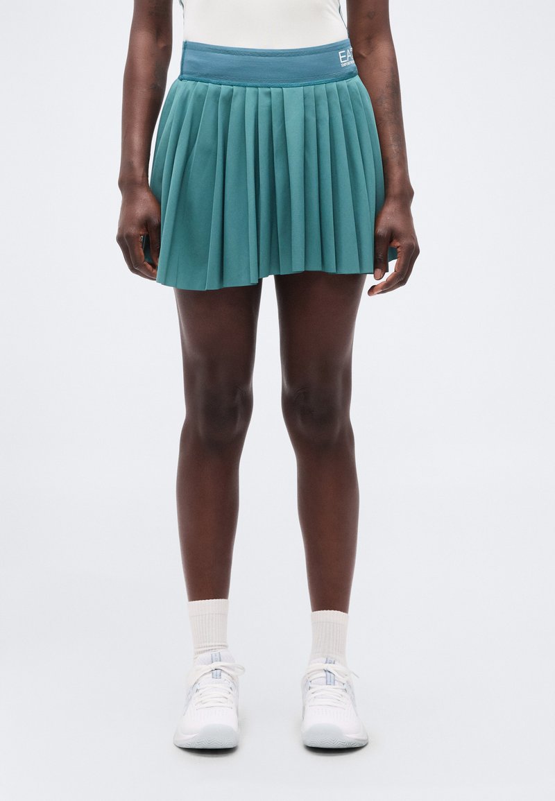 Άτομο που φοράει φούστα σε χρώμα τυρκουάζ με πιέτες, λευκά αθλητικά παπούτσια και λευκές κάλτσες, στέκεται μπροστά σε απλό ανοιχτό φόντο.