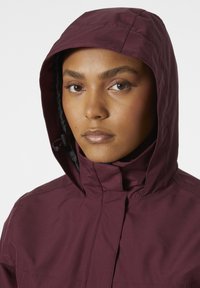 Chaqueta con capucha color burdeos y cuello alto, fabricada con un material suave y resistente al agua. Presenta cordones en la capucha para ajustar.