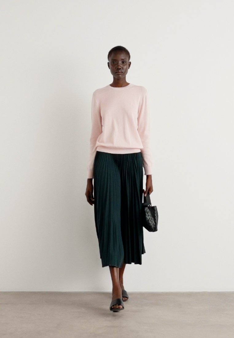 Pull en tricot rose clair avec un col rond, associé à une jupe plissée de longueur sol en vert foncé. Tenant un sac à main noir texturé.
