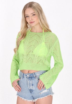 Pulover scurt tricotat de culoare verde neon, cu un model în zigzag și mâneci tip clopot, asortat cu pantaloni scurți din denim distresat, cu margini franjurate.