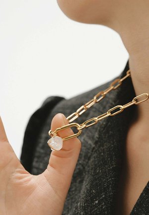 Persona sosteniendo un collar de cadena de oro con eslabones grandes y un colgante translúcido de forma irregular sobre una tela oscura texturizada.