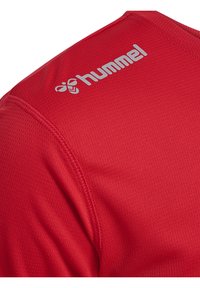 Rød sportsjersey med mesh tekstur, forstærket syning og sølvlogo på skulderen. Kortærmet, crew neck design.