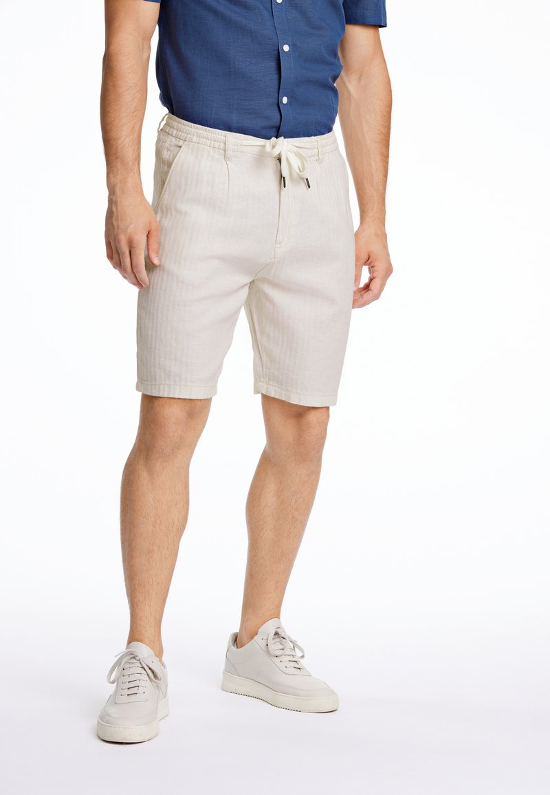 Pantalones cortos de lino beige con una cinturilla elástica y cordón ajustable. Combinados con zapatillas gris claro. El fondo es blanco liso.