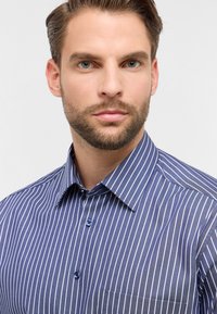 Camicia a righe blu scuro con linee verticali bianche. Presenta un colletto button-down, bottoni blu e una tasca sul petto. Tessuto dalla consistenza liscia.