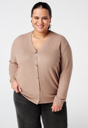Cardigan beige en V en tissu doux, avec des boutons dorés et des manches longues. Associé à un pantalon gris foncé.
