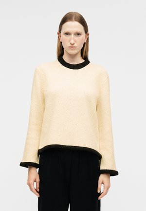 Kvinde med lyst brunt, glat hår iført en cremefarvet sweater med sort kant ved kraven, ærmerne og kanten, kombineret med sorte bukser, står neutralt.