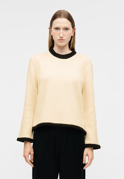 Femme aux cheveux châtain clair et raides portant un pull crème avec un bord noir au col, aux poignets et à l'ourlet, associé à un pantalon noir, en position neutre.