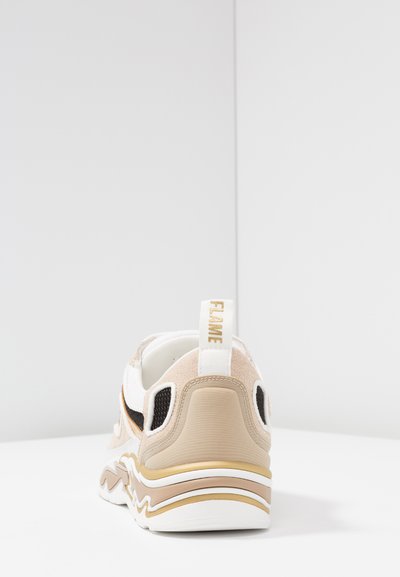 Sneaker med en beige och vit ovandel med svarta accenter, nätdetaljer och en guldrandig sula; inkluderar en ögla på hälen märkt 'FLAME.'