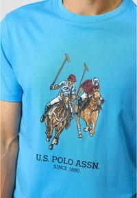 Šviesiai mėlynas medvilninis marškinėlis su grafika, vaizduojančia du polo žaidėjus ant žirgo, žemiau yra užrašas "U.S. POLO ASSN. SINCE 1890".