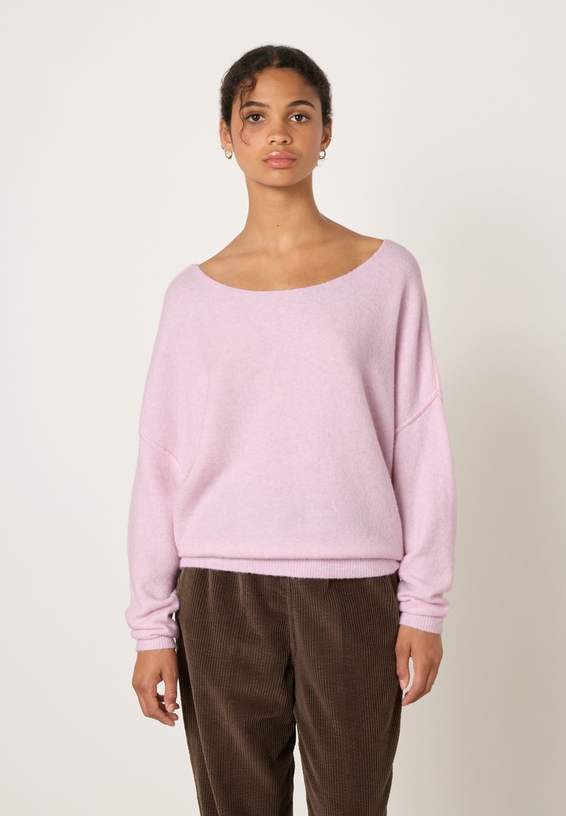 Jauna moteris dėvi laisvos formos šviesiai rožinį megztinį ir rudus kordurojus kelnes, stovinti priešais paprastą šviesų foną.