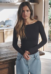 Mustast triiksärgist pikkade varrukatega, paadikaelusega, kombineeritud hele siniste kõrge vöökohaga teksadega, kantud loomulikus siseolekus.