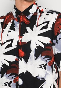 Camicia a maniche corte con un motivo floreale nero, bianco e rosso, realizzata in tessuto leggero, con colletto classico e chiusura a bottoni.