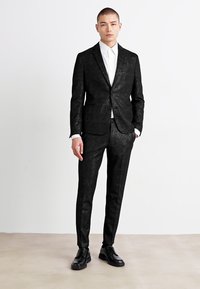 Fekete, szabott öltöny texturált felülettel, egy gombos zakóval és slim fit nadrággal. Fehér inggel és fekete cipővel párosítva.