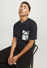 Sort bomulds t-shirt med rund hals og korte ærmer, der har en hvid mønstret lomme på venstre bryst. Sorte bukser.