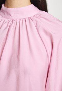 Blusa rosa con collo alto, dettagli arricciati sulle spalle e una consistenza del tessuto morbida e liscia.
