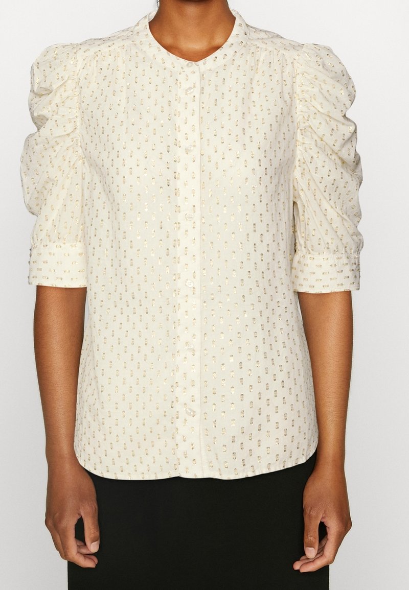 Blouse crème à manches 3/4 bouffantes avec motif de pois dorés, boutonnée devant, portée avec une jupe noire.