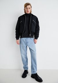 Chaqueta bomber negra con cierre delantero y dos bolsillos en el pecho, usada sobre un suéter de cuello alto oscuro. Jeans de corte relajado en azul claro y zapatos negros.