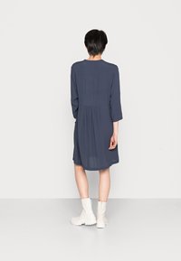 Kvinna med kort mörkt hår som bär en lös, långärmad marinblå klänning och vita ankelstövlar, stående vänd bort mot en enfärgad bakgrund.