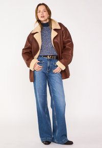 Veste en cuir marron avec col en shearling tan, pull en maille bleu et jeans en denim à jambes larges. Des bottes noires et une ceinture marron viennent compléter le look.