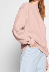 Różowa bluza z okrągłym dekoltem i długimi rękawami, z mankietami i luźnym krojem, zestawiona z jasnoniebieskimi dżinsami.