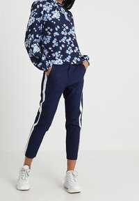 Blouse florale marine avec des fleurs bleu clair, manches longues et col à revers, associée à un pantalon fuselé marine avec des bandes blanches sur les côtés.