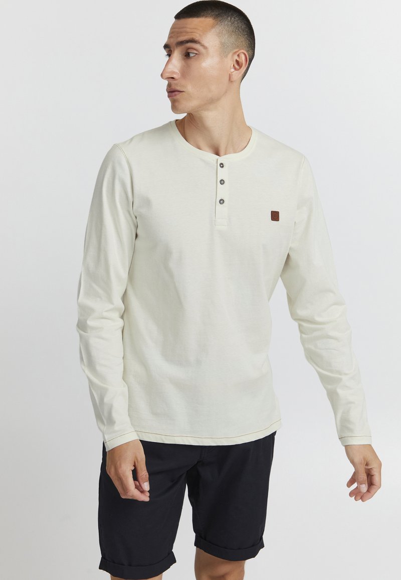 Camisa de manga larga color crema con cuello henley, tapeta con tres botones y un pequeño parche de logo marrón en el pecho; combinada con pantalones cortos negros.