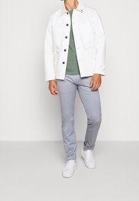 Bílá plátěná bunda s hnědými knoflíky, zelené pruhované tričko, světle šedé chinos a bílé tenisky. Jednoduchý design s praktickými kapsami.