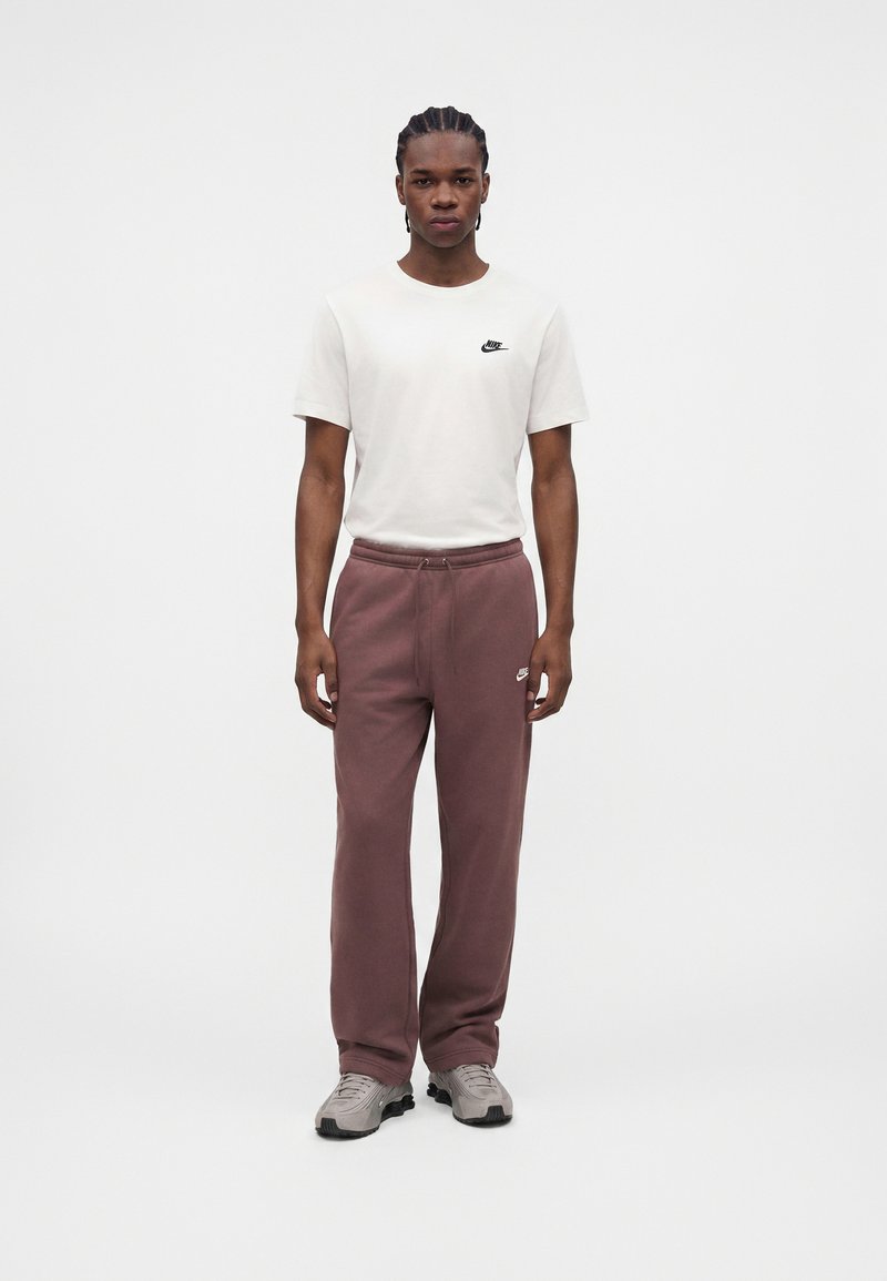 Junger Mann mit Zöpfen trägt ein weißes Nike-T-Shirt, mauve Nike-Sweatpants und graue Sneaker, steht vor einem schlichten weißen Hintergrund.