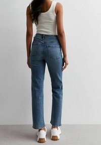 Kvinna står vänd bort, iklädd en vit linne, blå raka jeans och vita sneakers med gummisulor.