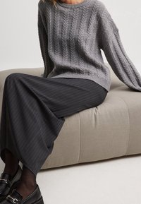 Grau gestrickter Pullover mit lockerer Passform, kombiniert mit einem langen, dunkelgrauen, gestreiften Rock. Schwarze Schuhe, sitzen auf einem beigen, runden Hocker.