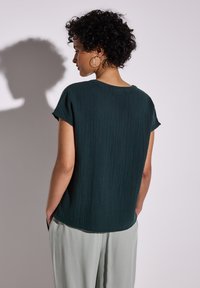 Top corto a maniche corte di un verde scuro e texture, visto da dietro, abbinato a pantaloni grigio chiaro. Tasche laterali visibili.