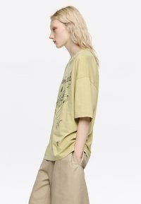 Camiseta de gran tamaño en verde oliva con diseño gráfico negro, mangas cortas y cuello redondo. Combinada con pantalones holgados beige.