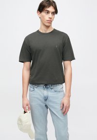 Σκούρο πράσινο κοντομάνικο μπλουζάκι με στρογγυλή λαιμόκοψη, χαλαρή εφαρμογή και μικρό λογότυπο στο στήθος, σε συνδυασμό με ανοιχτόχρωμο μπλε τζιν και μπεζ καπέλο.