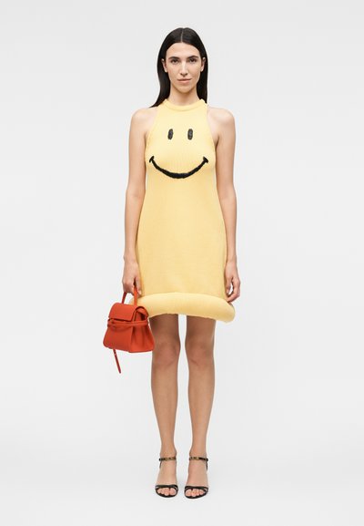 Γυναίκα με κίτρινο φόρεμα χωρίς μανίκια με μαύρο σχέδιο χαμογελαστής φατσούλας, κρατάει μικρή κόκκινη τσάντα και φοράει μαύρα πέδιλα με λουράκια.
