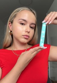 SWEED-produkt i en mintgrønn tube med sky-mønster; holdt nær en person iført rød skjorte og gull hoop-øredobber.