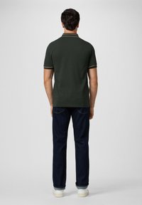 Man in een donkergroene polo met korte mouwen en beige biezen, donkere spijkerbroek en witte sneakers, staand met de rug naar de camera tegen een grijze achtergrond.