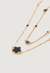 Collar de cadena de oro con colgantes en forma de trébol negro y pequeños acentos de gema negra sobre un fondo claro.