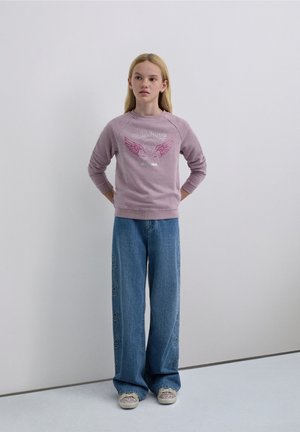 Sudadera de color púrpura claro con estampado gráfico, combinada con pantalones vaqueros de pierna ancha azul con bordado de estrellas. Zapatillas deportivas casuales con detalles de purpurina.