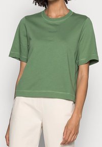 Persona con una semplice maglietta verde a maniche corte e pantaloni crema, in piedi con un braccio rilassato lungo il fianco.