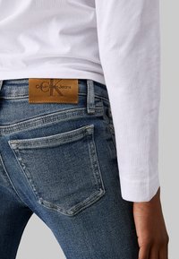 Vaqueros de mezclilla azul oscuro con ligero desgaste, que presentan un parche de cuero marrón con la etiqueta "Calvin Klein Jeans" y bolsillos traseros visibles. Camisa de manga larga blanca.