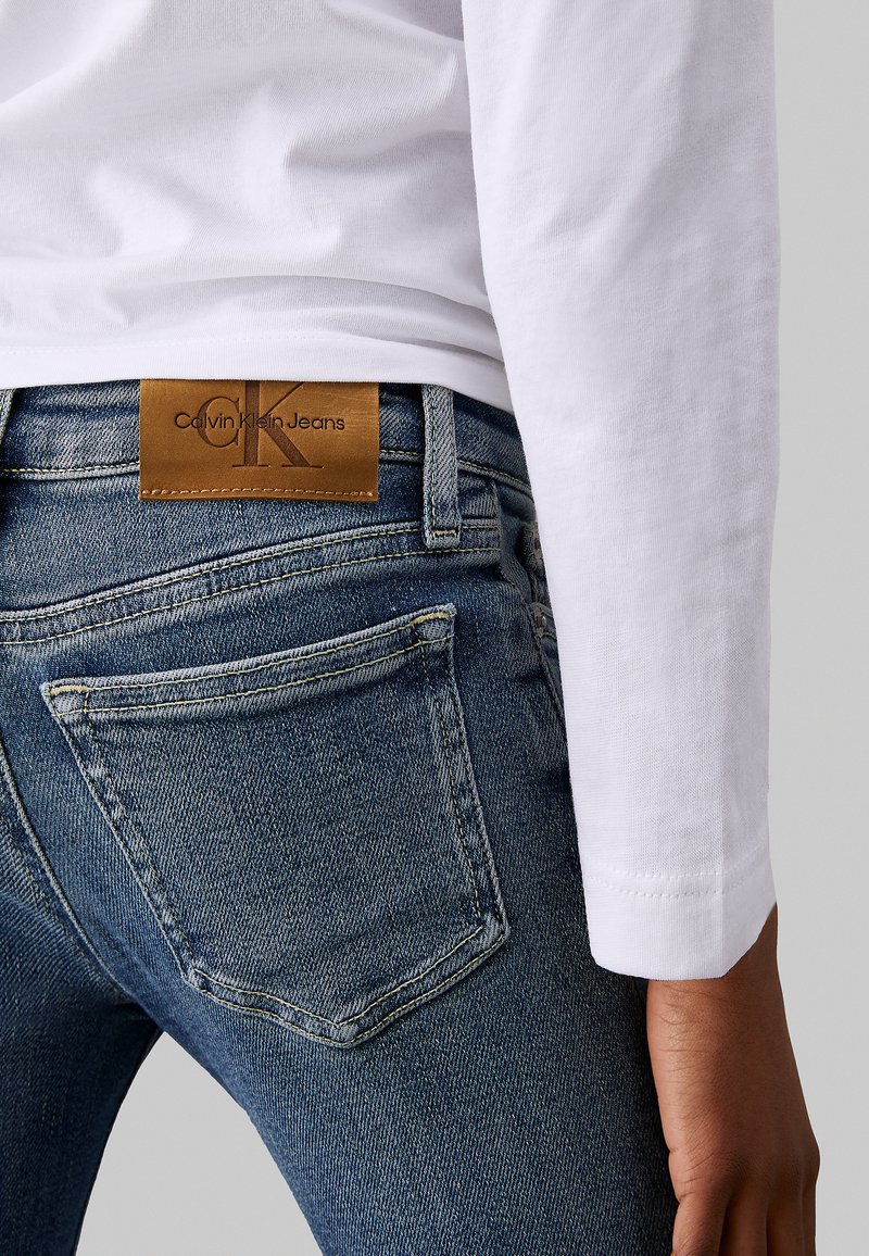 Dunkelblaue Jeans aus Denim mit leichter Abnutzung, versehen mit einem braunen Lederpatch mit der Aufschrift "Calvin Klein Jeans" und sichtbaren Gesäßtaschen. Weißes Langarmshirt.