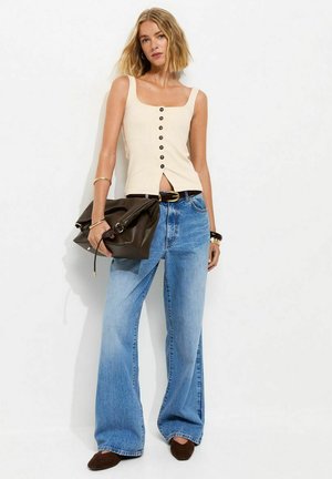 Ung kvinde iført beige knappet tank top, vidde blå jeans, brunt bælte, holder en stor brun clutch, med kort blondt hår.