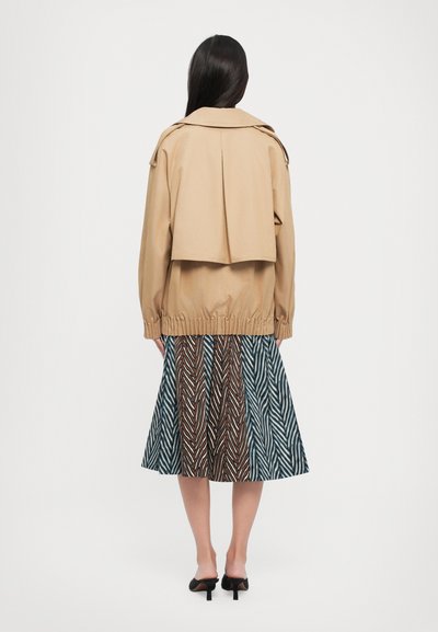 Beige jasje met een cropped ontwerp en geplooide achterkant, gecombineerd met een multi-kleurige patroonrok met zigzag- en diagonale lijnen.