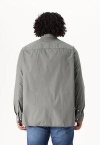 Camisa gris de botones con mangas largas, corte recto y ligero brillo; vista trasera que muestra un diseño liso y sin patrones ni acentos visibles.