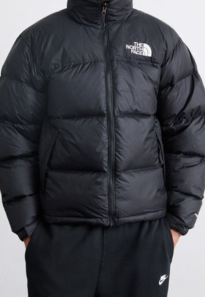 Doudoune noire avec fermeture éclair à l'avant et logo The North Face portée par une personne avec les mains dans les poches d'un pantalon de survêtement Nike noir.